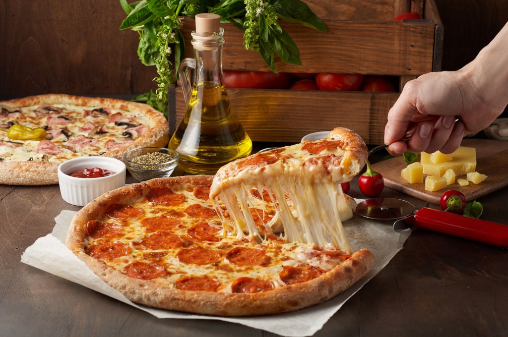 The nearby appearance of a female hand holding a slice of hot pizza with molten cheese and chorizo on a rustic wooden table, surrounded by ingredients
