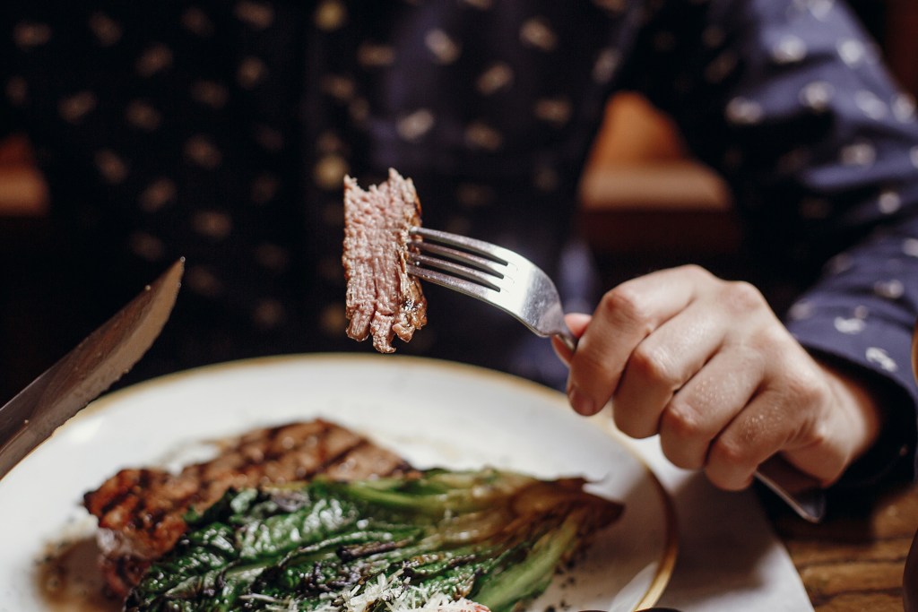 The person who uses a fork and knife to eat a steak with grilled vegetables.