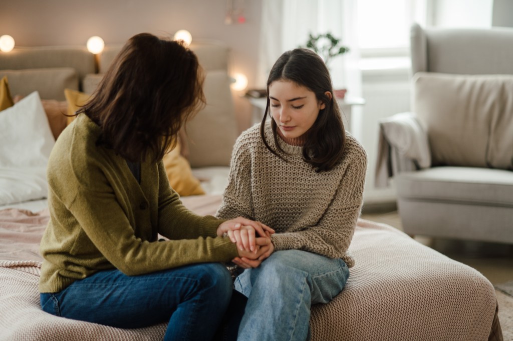 A teenage mother and girl holding her hands and talking on a bed.