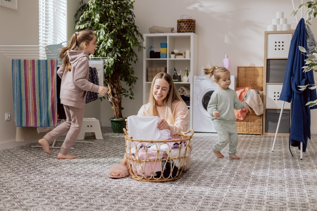 A mother order laundry in a basket while her two young daughters play nearby.